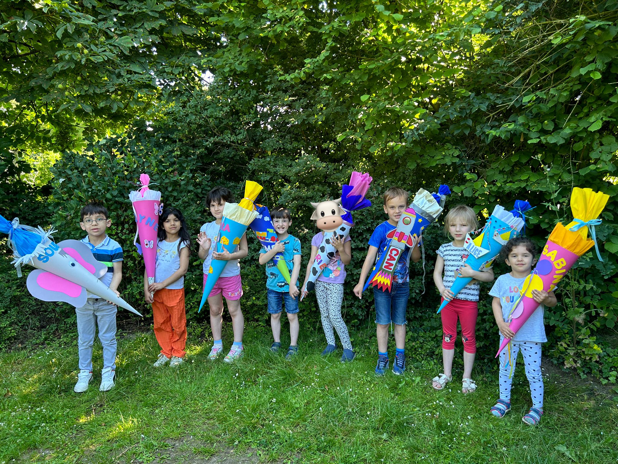Acht Kinder mit ihren bunten Schultüten stehen vor einem Busch.