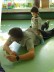 Ein Lehrer liegt mit Sportkleidung auf dem Bauch. 
Auf seinem Rücken sitzt ein Junge.