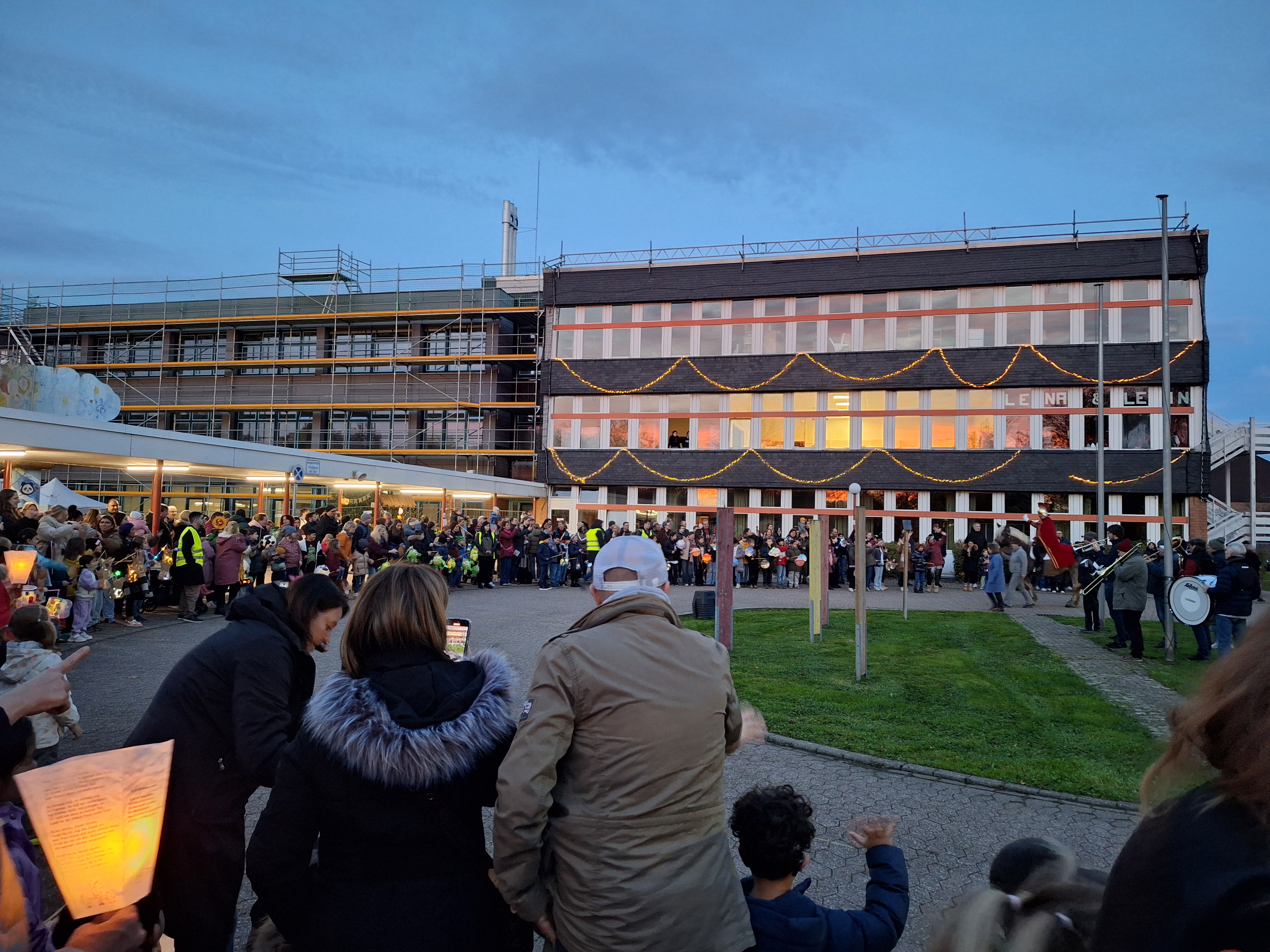 sehr viele Kinder mit Laternen und Erwachsene stehen am Rondell der Schule, Sankt Martin zieht auf einem Pferd ein