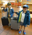 2 Jungen stehen mit ihren Koffern, Sonnenbrillen und Jacken im Kindergarten