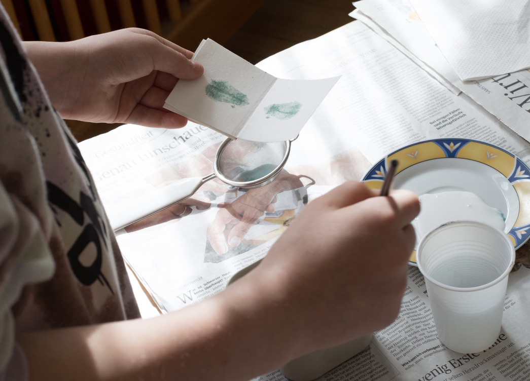 Man sieht Kinderhände, die eine Faltkarte halten. Auf dem Bild ist ein Tisch mit Wasserbecher, Pinsel und Papier.