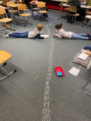 Zwei Kinder liegen in einem Klassenraum auf dem Boden und legen Mengen mit Wattestäbchen.