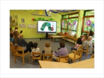 Kindergartenkinder und Erzieher sitzen im Halbkreis, sie schauen auf ein projiziertes Bild der Raupe Nimmersatt