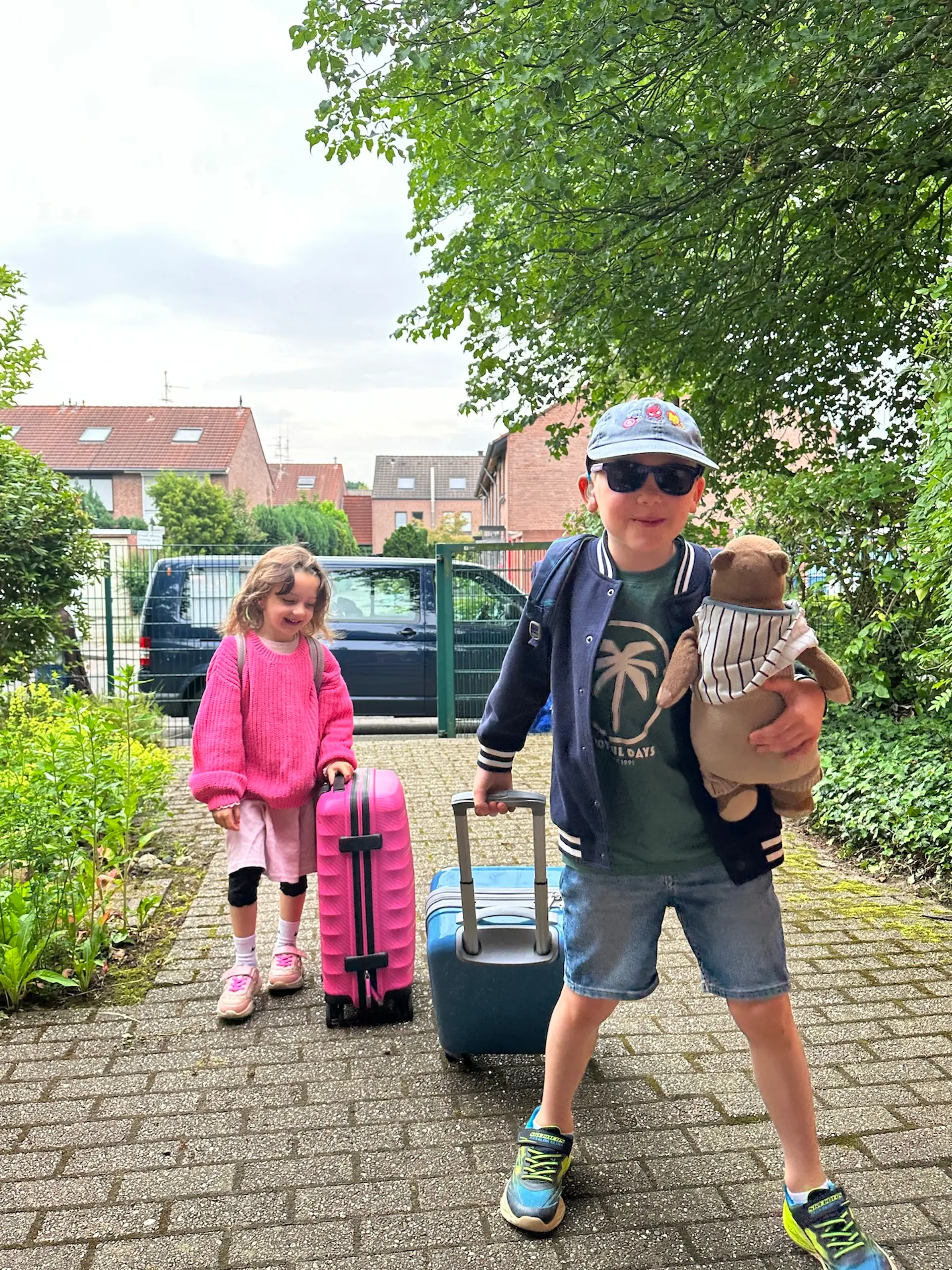 2 Kinder mit Rollkoffern, eines davon hält ein Kuscheltier in der Hand 