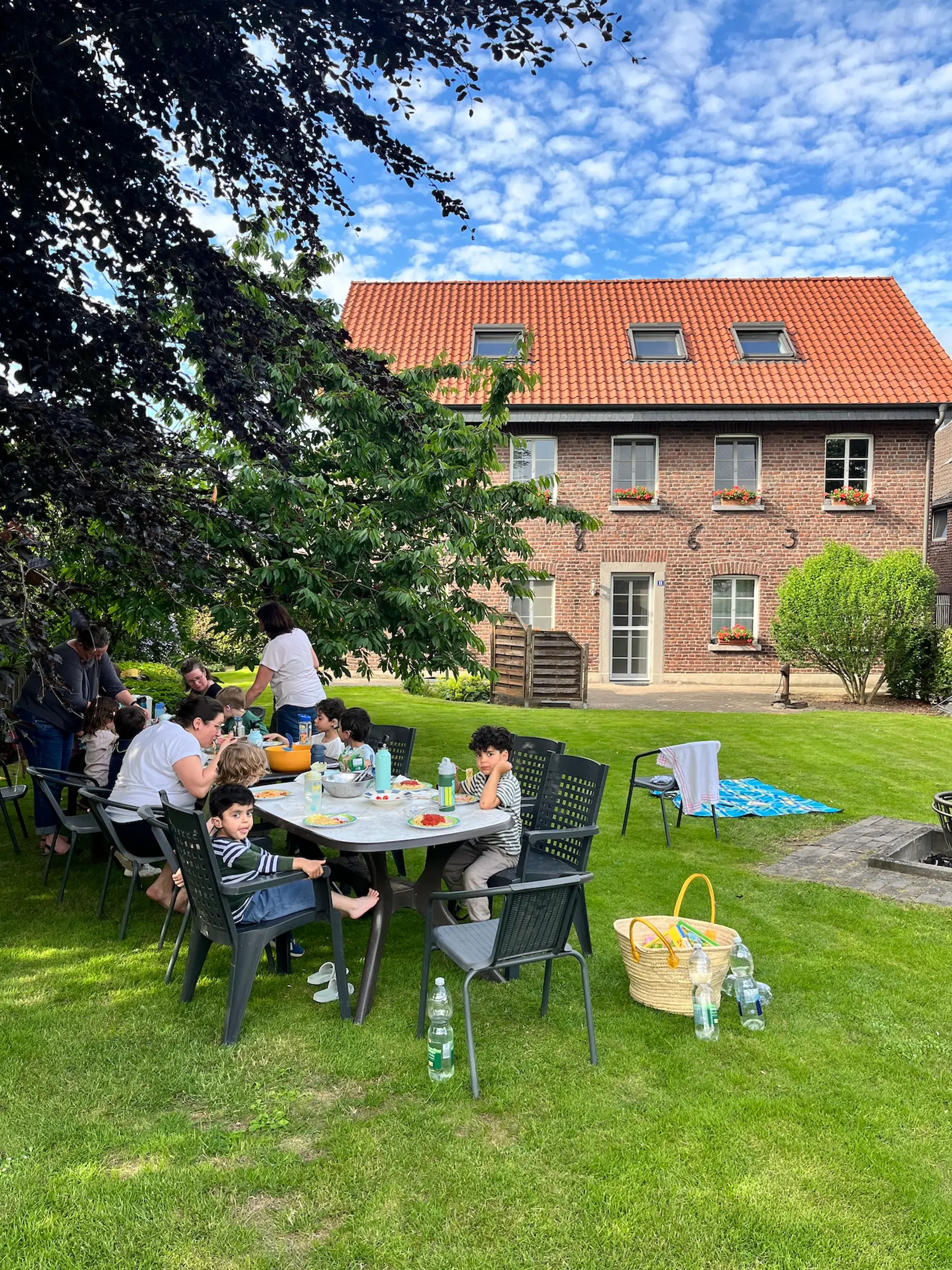 Auf einer Wiese vor einem Bauernhaus sitzen Kinder und Lehrerinnen an einem langen Tisch