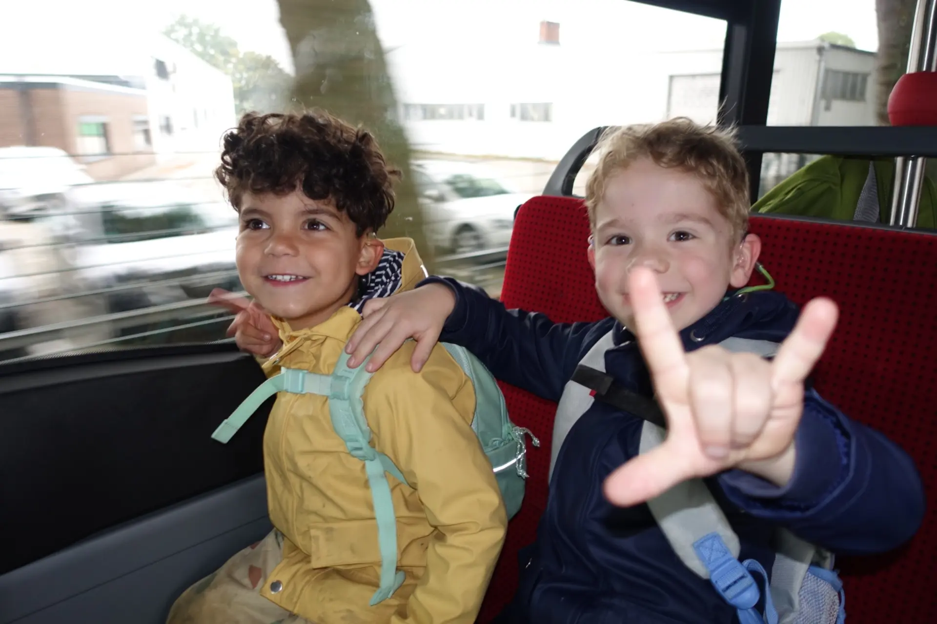 Zwei Kindergartenkinder sitzen im Bus und winken fröhlich in die Kamera