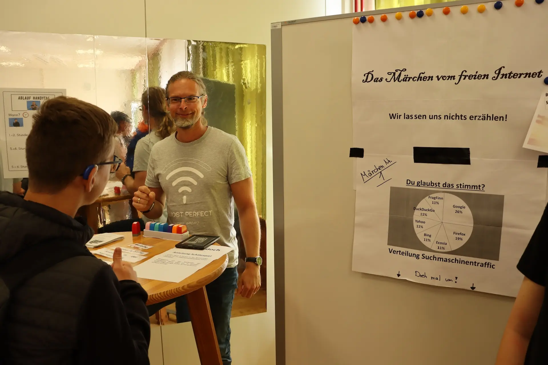 Ein Lehrer steht neben einer Stellwand zum Thema "Das Märchen vom freien Internet" und spricht mit einem Schüler, den man von hinten sieht