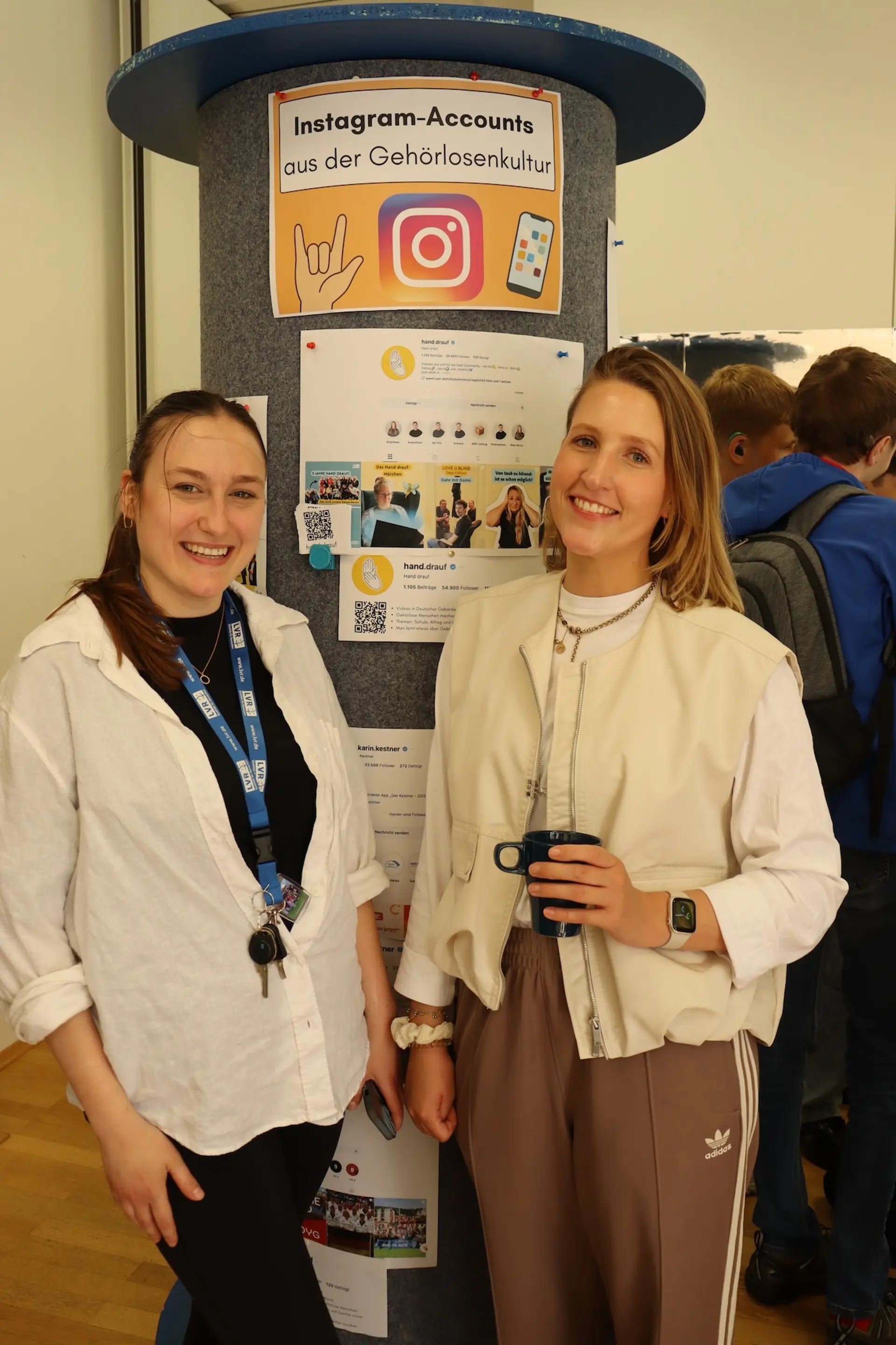zwei Lehrerinnen stehen vor einer Litfass-Säule zum Thema "Instagram-Accounts aus der Gehörlosenkultur"