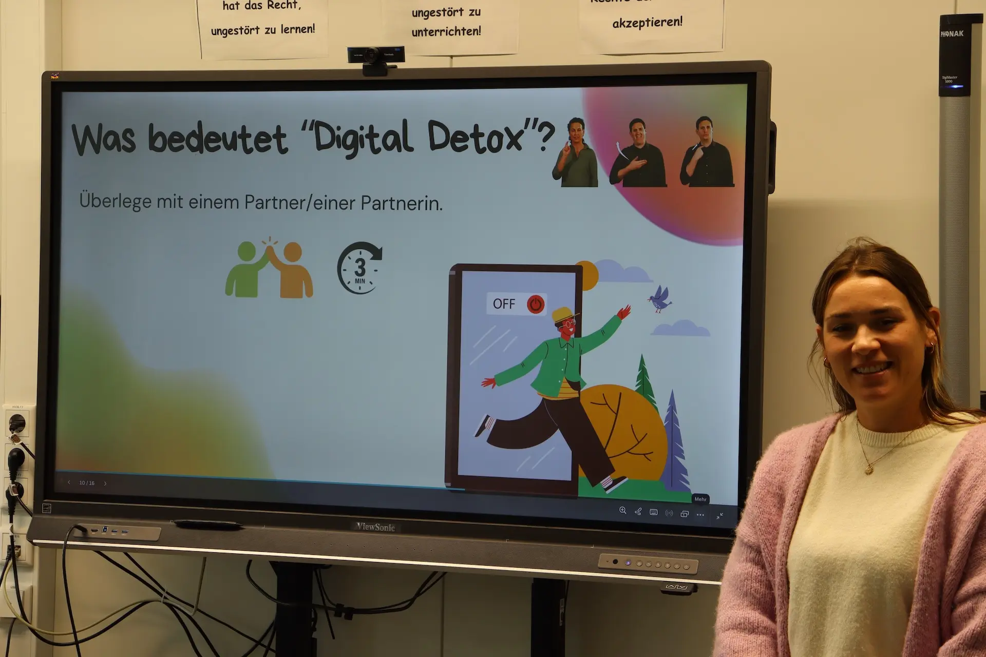 eine Lehrerin steht neben einer digitalen Tafel mit der Überschrift "Was bedeutet Digital Detox?"