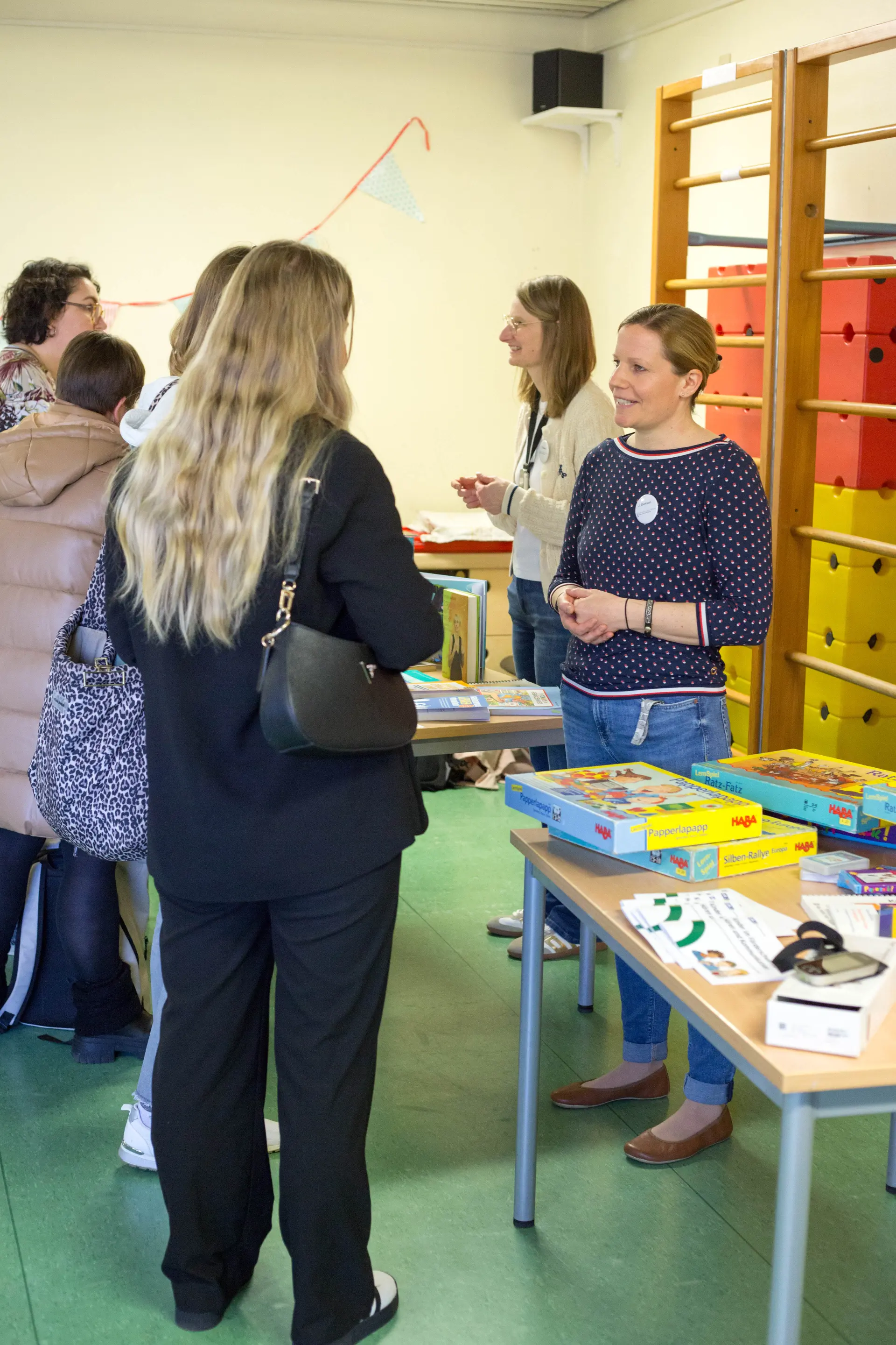 im Bewegungsraum schauen sich Erzieher*innen (von der Seite zu sehen) Infomaterial auf Tischen an und sprechen mit den Lehrer*innen