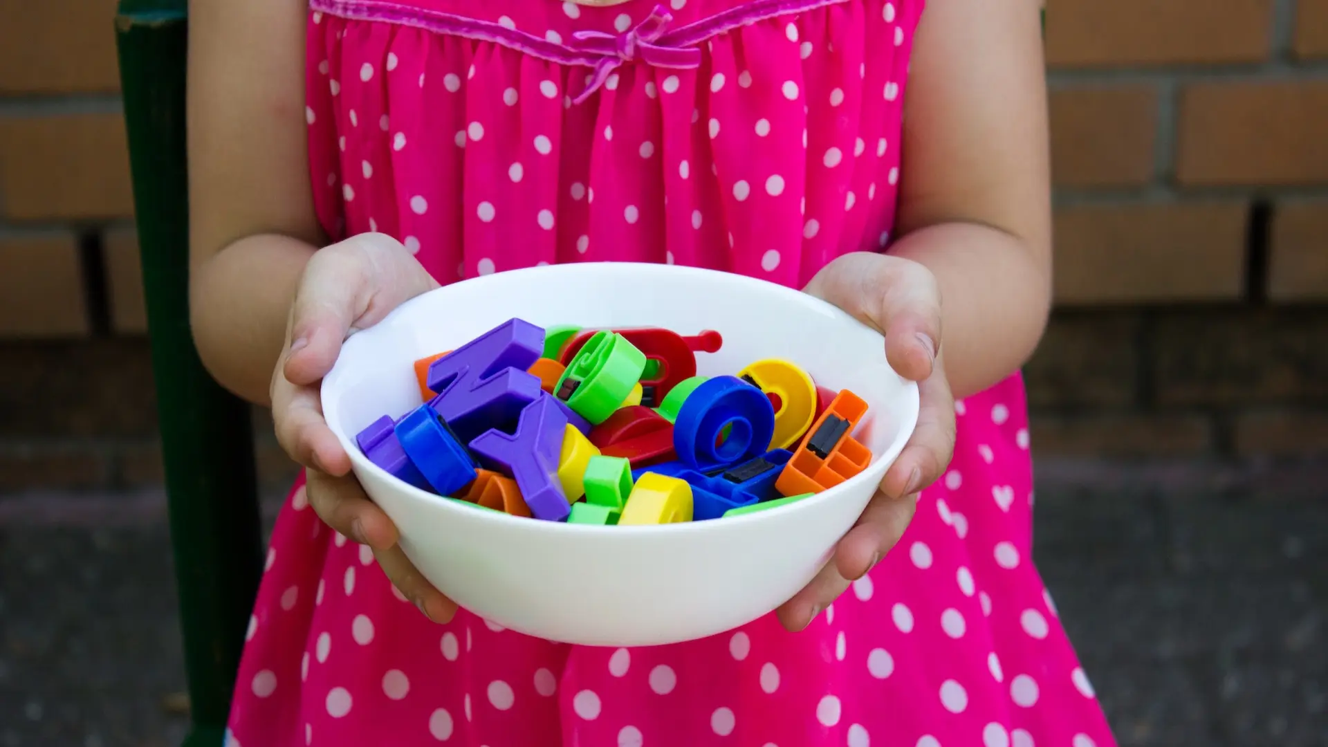 Kind im rosa Kleid hält eine Schüssel mit bunten Buchstaben aus Plastik in die Kamera