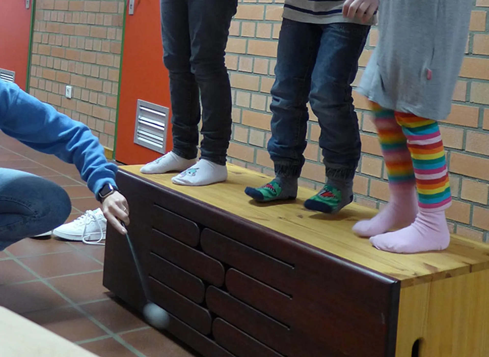Drei Kinder stehen auf einer Sportbank und eine Lehrkraft schlägt mit einem Trommelstock auf die Seite, um Schallwellen spürbar zu machen